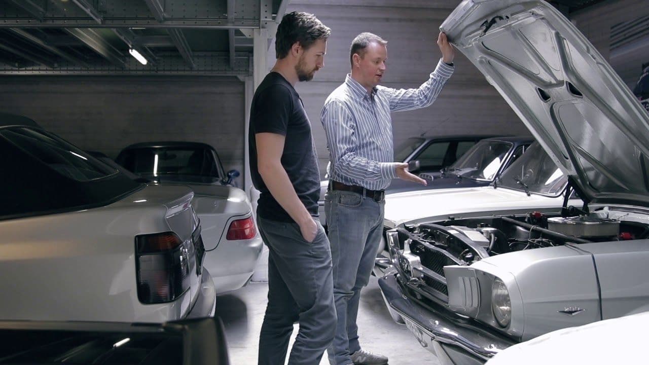 Two people examine the engine of a classic car with its bonnet open.
